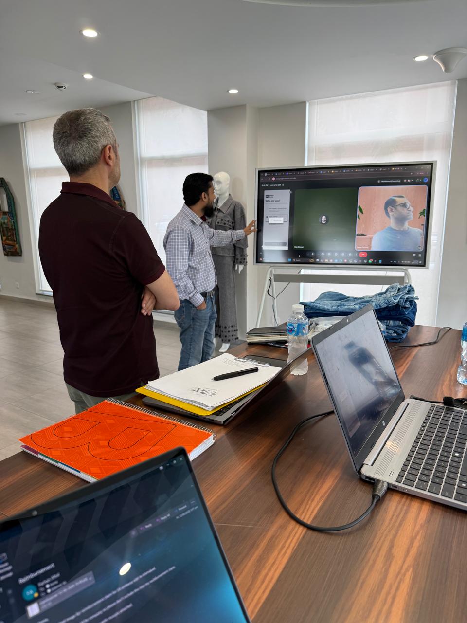 DenimNotes live platform demo running on Diamond Denim's large display screen — team member presenting to in-person and remote participants while collection and garments are visible in background