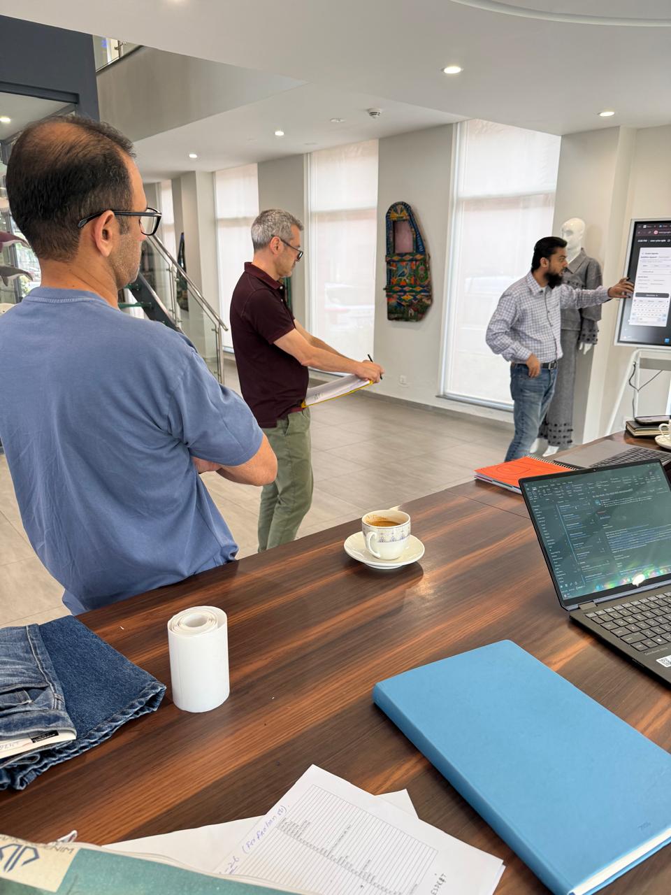 Diamond Denim showroom — team reviewing DenimNotes platform on screen with garment collection and mannequin in background showing grey denim coordinates